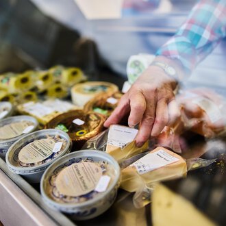 © Kleinwalsertal Tourismus | Fotograf Oliver Farys Ein Verkaufsstand mit verschiedenen Käsesorten. Eine Hand greift nach einem Käseprodukt in der Auslage. | © Kleinwalsertal Tourismus | Fotograf Oliver Farys