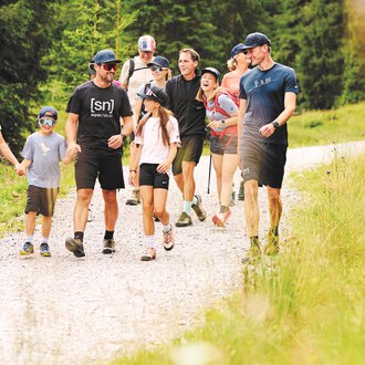 Eine Gruppe von Menschen wandert auf einem Schotterweg durch eine grüne Landschaft. Umgeben von Bäumen genießen sie die Natur zusammen. | © Kleinwalsertal Tourismus | Oliver Farys