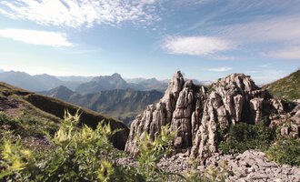 Eine beeindruckende Berglandschaft mit schroffen Felsen und majestätischen Bergen im Hintergrund. Der Himmel ist klar und die Umgebung ist von grünem Pflanzenbewuchs geprägt. | © Kleinwalsertal Tourismus eGen | Frank Drechsel