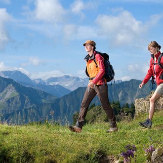 Zwei Wanderer gehen durch eine grüne Landschaft mit Bergen im Hintergrund. Der Himmel ist blau mit einigen Wolken. | © Kleinwalsertal Tourismus | Andi Mayr