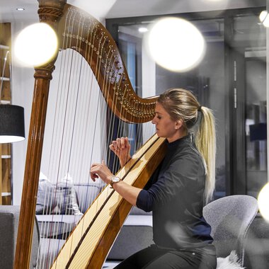 Eine Frau spielt Harfe in einem modernen Raum. Weiches Licht und stilvolle Möbel schaffen eine gemütliche Atmosphäre. | © Genuss- & Aktivhotel Sonnenburg | Michael Gunz