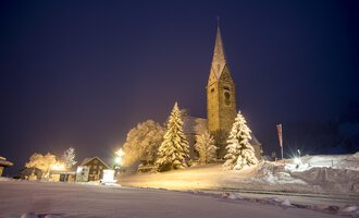 © Kleinwalsertal Tourismus | Dominik Berchtold A snowy landscape at night with a church and illuminated trees. The sky is dark blue and the road is visible. | © Kleinwalsertal Tourismus | Dominik Berchtold