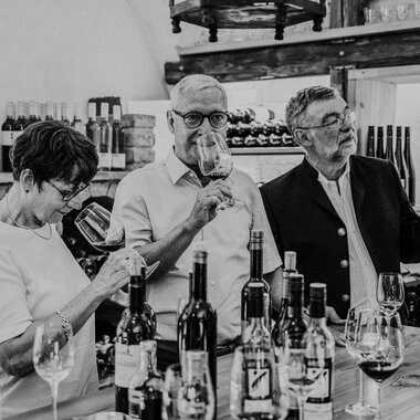 A group of three people is enjoying wine in a wine shop. They are holding glasses in their hands and are looking intently at the glasses. | © Das Naturhotel Chesa Valisa
