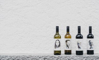 Vier Flaschen Wein stehen auf einer grauen Fläche. Die Etiketten zeigen schwarz-weiße Porträts von verschiedenen Personen. | © Das Naturhotel Chesa Valisa
