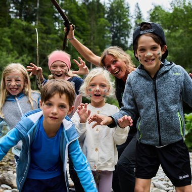 Eine Gruppe von Kindern und einem Erwachsenen, die am Ufer eines Flusses stehen. Sie haben Spaß und posieren mit fröhlichen Gesichtsausdrücken. | © Kleinwalsertal Tourismus | Oliver Farys