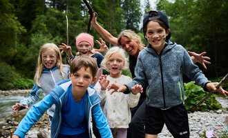 Eine Gruppe von Kindern und einem Erwachsenen, die am Ufer eines Flusses stehen. Sie haben Spaß und posieren mit fröhlichen Gesichtsausdrücken. | © Kleinwalsertal Tourismus | Oliver Farys