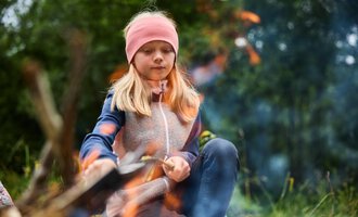 Ein Mädchen sitzt am Lagerfeuer und schaut konzentriert in die Flammen. Der Hintergrund zeigt eine grüne Wiesenlandschaft. | © Kleinwalsertal Tourismus | Oliver Farys