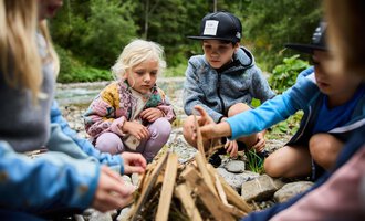 Eine Gruppe von Kindern versammelt sich um ein kleines Lagerfeuer. Sie sind von Natur umgeben und konzentrieren sich auf das Feuermachen. | © Kleinwalsertal Tourismus | Oliver Farys