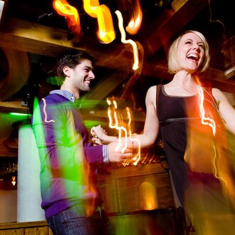 © Aparthotel Kleinwalsertal Haller GmbH & Co KG A couple dances happily in a lively club. Colorful lights and a festive atmosphere surround them. | © Aparthotel Kleinwalsertal Haller GmbH & Co KG