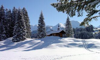 A snowy landscape with tall fir trees and a small wooden house. In the background, the snow-covered mountains and a clear blue sky can be seen. | © Bruder Klaus Kapelle | Rolf Koeberle