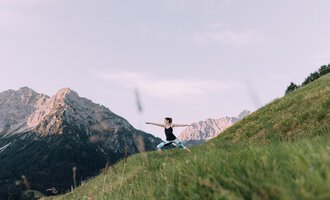 A person is practicing yoga in nature on a green meadow. Impressive mountains are visible in the background. | © Naturhotel Chesa Valisa