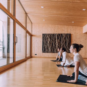 © Naturhotel Chesa Valisa Ein heller Yogaraum mit Holzverkleidung und großen Fenstern. Drei Personen praktizieren Yoga und blicken nach draußen. | © Naturhotel Chesa Valisa