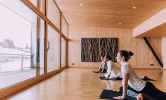 Ein heller Yogaraum mit Holzverkleidung und großen Fenstern. Drei Personen praktizieren Yoga und blicken nach draußen. | © Naturhotel Chesa Valisa