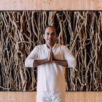A man stands with his hands folded in front of a wall made of natural materials. He is wearing a simple, light outfit and exudes calmness. | © Das Naturhotel Chesa Valisa