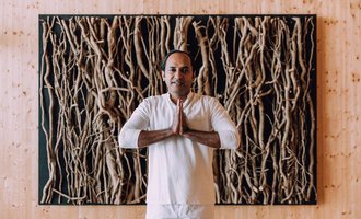 A man stands with his hands folded in front of a wall made of natural materials. He is wearing a simple, light outfit and exudes calmness. | © Das Naturhotel Chesa Valisa