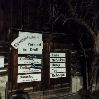 A wooden building with a sign for specialty sales in the barn. The signage indicates opening hours and offered products such as cheese and honey. | © Abler Spezialitätenverkauf im Stall | N. Lughammer
