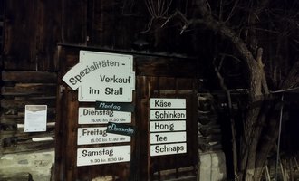 A wooden building with a sign for specialty sales in the barn. The signage indicates opening hours and offered products such as cheese and honey. | © Abler Spezialitätenverkauf im Stall | N. Lughammer