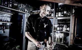 A bartender working at a well-stocked bar. He smiles while he is busy with a glass. | © Verwöhn- und Wellnesshotel Walserhof | Werner Krug