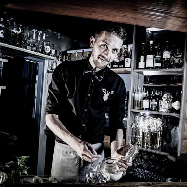 A bartender working at a well-stocked bar. He smiles while he is busy with a glass. | © Verwöhn- und Wellnesshotel Walserhof | Werner Krug