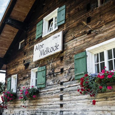 Ein traditionelles Holzhaus mit grünen Fensterläden. Blumenkästen mit bunten Blumen schmücken die Fenster. | © Kleinwalsertal Tourismus | Andre Tappe