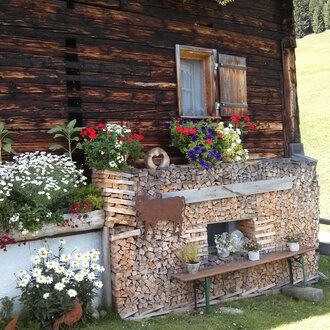 Ein traditionelles Holzhaus mit bunten Blumen und Holzstapel. Der Ort strahlt ländliche Gemütlichkeit und Naturverbundenheit aus. | © Kleinwalsertal Tourismus | N. Lughammer
