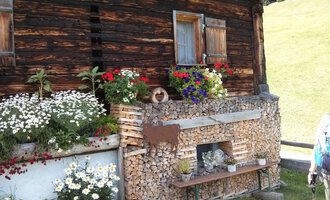 Ein traditionelles Holzhaus mit bunten Blumen und Holzstapel. Der Ort strahlt ländliche Gemütlichkeit und Naturverbundenheit aus. | © Kleinwalsertal Tourismus | N. Lughammer