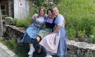 Drei Frauen sitzen auf einer Steinmauer in traditioneller Tracht. Im Hintergrund ist eine grüne Wiese und ein Holzhaus zu sehen. | © Alpe Widderstein | Tom Egger
