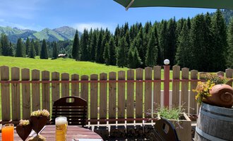 A cozy outdoor area with a table and drinks. In the background, there is a beautiful green landscape with forest and mountains. | © Alpenhotel DAS KÜREN