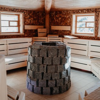 A cozy sauna room with wooden benches and large windows. In the center stands a stone oven. | © Verwöhn- & Wellnesshotel Walserhof| Aileen Melucci
