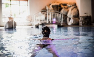 A woman is swimming in a relaxing indoor pool. In the background, rocks and gentle lighting can be seen. | © Verwöhn- & Wellnesshotel Walserhof| Aileen Melucci