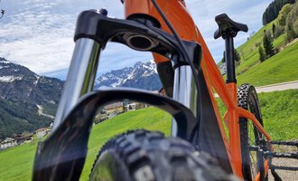An orange mountain bike in a green landscape with mountains in the background. The sky is slightly cloudy, and it is an idyllic setting. | © Austrian Ski- & Service Ranch | Trixi Leitner