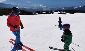 Two children are standing on skis in a sunny ski resort. In the background, snow-covered mountains and a clear landscape can be seen. | © Austrian Ski- & Service Ranch | Trixi Leitner