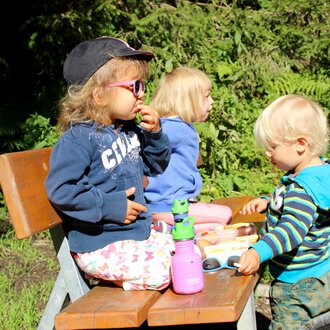 Three children are sitting on a bench outside. They are enjoying snacks and playing together. | © Kleinwalsertal Tourismus | Britta Maier