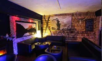 Ein gemütlicher Innenraum mit dunklen Ledermöbeln und einer Holzverkleidung. An der Wand hängt ein Bild eines Bergpanoramas und ein stilisiertes Ziegenmotiv. | © Heiko Möller m&m bar