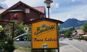 An inviting sign of the "Bura Lädele" in a picturesque landscape. In the background, mountains and a clear sky can be seen. | © Kleinwalsertal Tourismus | N. Lughammer