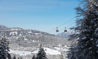 Eine Schneelandschaft mit schneebedeckten Bäumen und Bergen im Hintergrund. Zwei Gondeln schweben über die Winterlandschaft unter einem klaren blauen Himmel. | © OBERSTDORF · KLEINWALSERTAL BERGBAHNEN