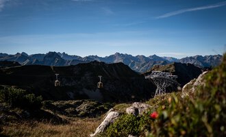 A cable car rides over a green meadow with rocks. In the background, majestic mountains and a clear sky can be seen. | © OBERSTDORF · KLEINWALSERTAL BERGBAHNEN