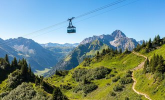 Eine Seilbahn schwebt über grüne Hügel und Berge. Der Himmel ist klar und die Landschaft ist malerisch. | © OBERSTDORF · KLEINWALSERTAL BERGBAHNEN