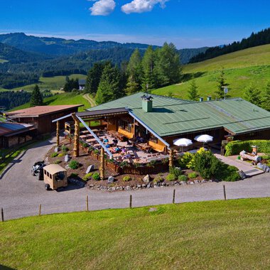 Eine malerische Almhütte umgeben von grünen Wiesen und Wald. Auf der Terrasse genießen Menschen die sonnige Aussicht in die Berge. | © Bergstüble | Jürgen Waffenschmidt