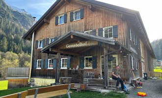 Ein traditionelles Holzhaus mit dem Namen "Gemstelhof" steht in einer ländlichen Umgebung. Vor dem Gebäude sitzen Bänke und ein Mensch ist sichtbar, während die Berge im Hintergrund zu sehen sind. | © Bernhards Gemstel Hofladen | Bernhard Heim
