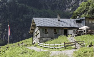 A cozy cabin in the mountains, surrounded by green meadows. In the foreground, a paved path leads to the front door, and a terrace with a sunshade is visible. | © Bernhards Gemstelalp | Frank Drechsel