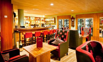 A cozy bar with a wooden interior and red chairs. The lighting is warm and inviting, with shelves full of drinks in the background. | © Hotel Birkenhöhe | Fotograf Krug