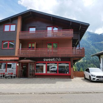 A modern building with a restaurant called "sweetchili" on the ground floor. In the background, green mountains and a blue sky can be seen. | © Kleinwalsertal Tourismus | N. Lughammer