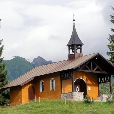 Eine kleine, orangefarbene Kirche steht umgeben von grünen Bäumen. Im Hintergrund sind Berge und eine bewölkte Himmel zu sehen. | © Bruder Klaus Kapelle | Monika Gmeiner
