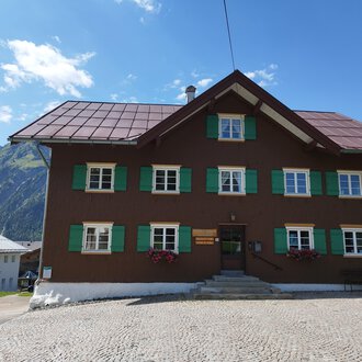 A traditional old house with a brown facade and green shutters. In the background, mountains and a blue sky are visible. | © Kleinwalsertal Tourismus | Nicole Lughammer