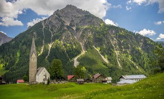A picturesque landscape with a village and an impressive mountain backdrop. In the foreground stands an old church and lush meadows. | © Kleinwalsertal Tourismus | Steffen Berschin