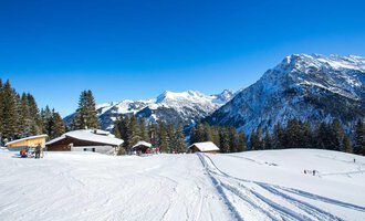 A snow-covered landscape with mountains and a clear blue sky. In the foreground, some huts and skiers can be seen. | © Bühlalpe | Frank Drechsel