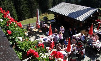 A cozy inn in the mountains with many guests outdoors. Red flowers and umbrellas create an inviting atmosphere in the surroundings. | © Bühlalpe | Frank Drechsel