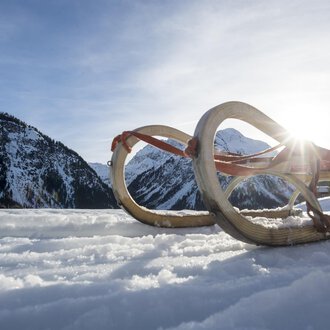 Ein Schlitten liegt im Schnee, umgeben von einer winterlichen Berglandschaft. Die Sonne strahlt hinter den Bergen hervor. | © Kleinwalsertal Tourismus | Dominik Berchtold