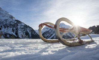 Ein Schlitten liegt im Schnee, umgeben von einer winterlichen Berglandschaft. Die Sonne strahlt hinter den Bergen hervor. | © Kleinwalsertal Tourismus | Dominik Berchtold